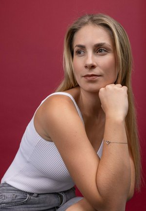 Silver chain bracelet with heart pendant, worn on a fair skin arm. Model in a white ribbed tank top against a solid maroon background.