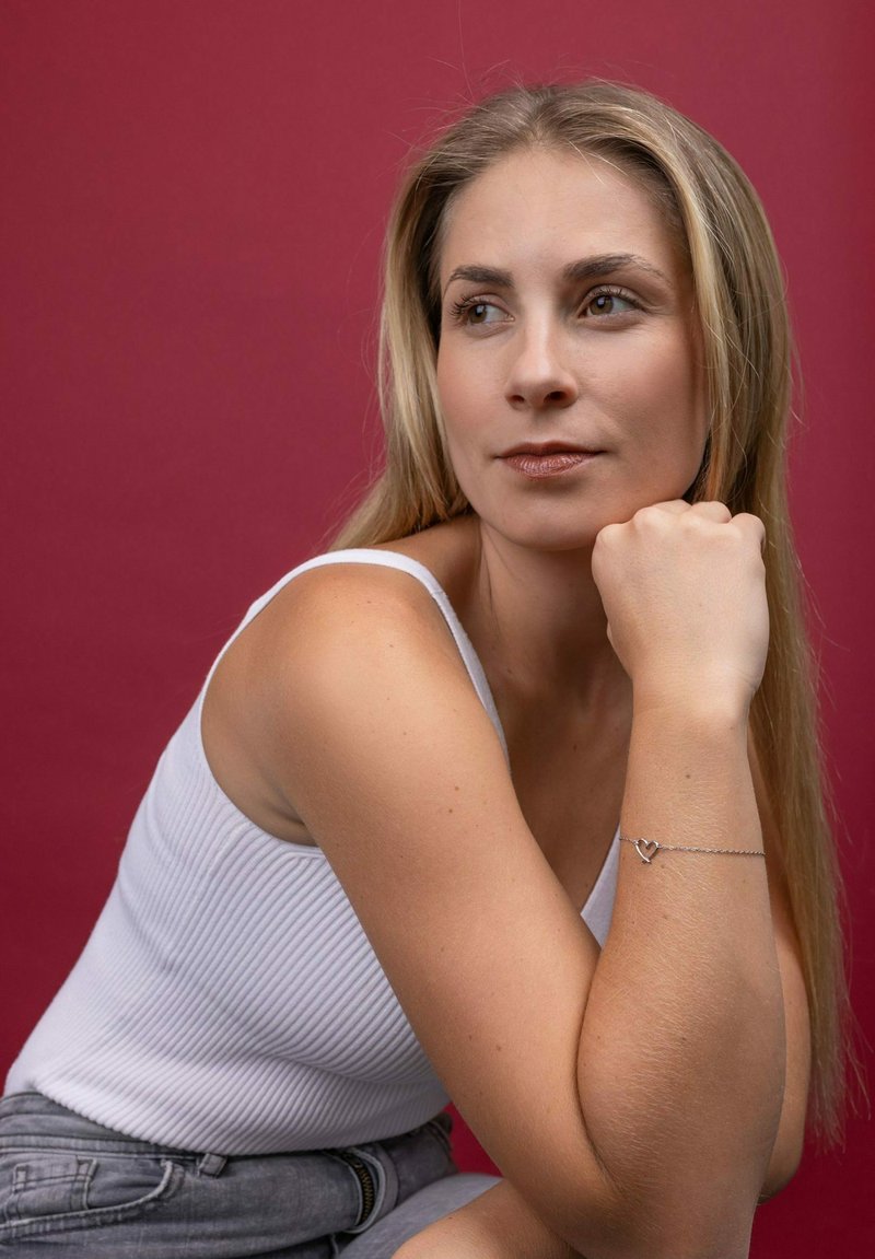 Stříbrný řetízkový náramek s přívěskem ve tvaru srdce, nošený na světlé kůži ruky. Modelka ve bílé žebrované tílku na jednobarevném tmavě červeném pozadí.