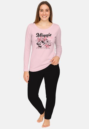 Giovane donna che sorride, indossando una maglia a maniche lunghe rosa chiaro con una grafica di Minnie Mouse e pantaloni neri, scalza su uno sfondo neutro.
