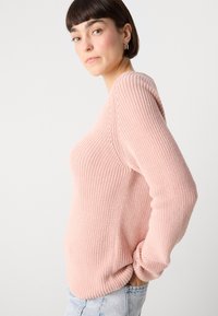 Šviesiai rožinė megzta džemperis su įpjovimu tekstūra, ilgomis rankovėmis ir laisvu siluetu, derinamas su šviesiai mėlynomis džinsinėmis kelnių.