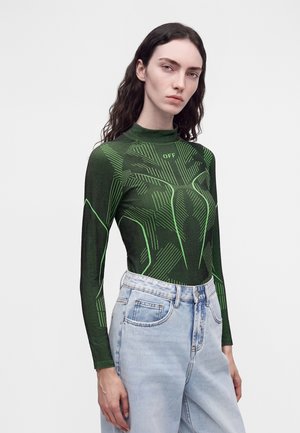 Mujer con cabello largo y oscuro que lleva una camiseta ajustada de color verde oscuro con líneas geométricas de color verde brillante y vaqueros de cintura alta de color azul claro.