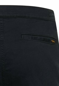 Black fabric pants showing a single back welt pocket with stitching details and a small rectangular black and gold label.