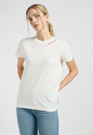 T-shirt bianco in cotone con maniche corte, con testo rosa sulla spalla. Design semplice e texture morbida, abbinato a jeans azzurri chiari.