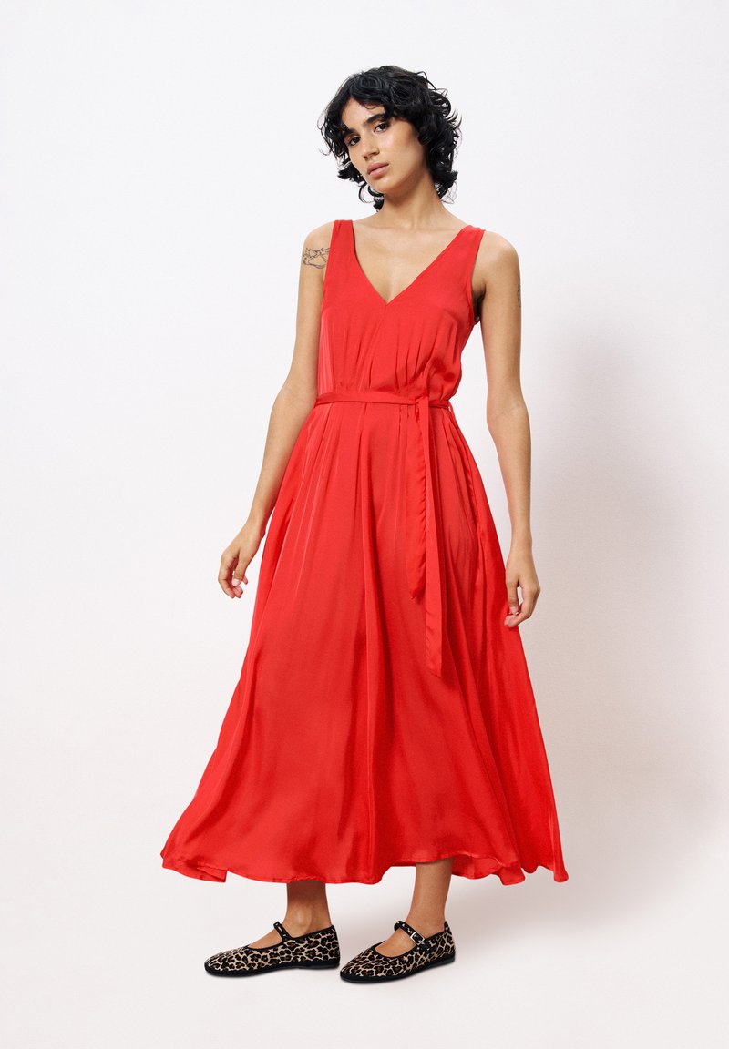 Red, sleeveless maxi dress in a flowing fabric with a V-neck and a tied waist. Worn with black and leopard print flats.