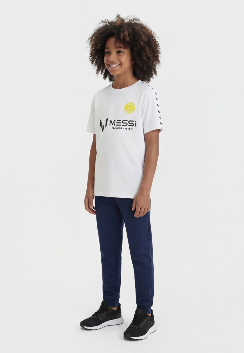 Mosolygós, göndör hajú gyermek fehér Messi Training System pólót, tengerészkék nadrágot és fekete sportcipőt viselve, fehér háttér előtt áll.