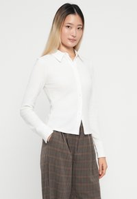 Bela rebrasta srajca z gumbi in ovratnikom ter dolgimi rokavi, ki ima ozek kroj. Model jo nosi z karo visokimi hlačami.