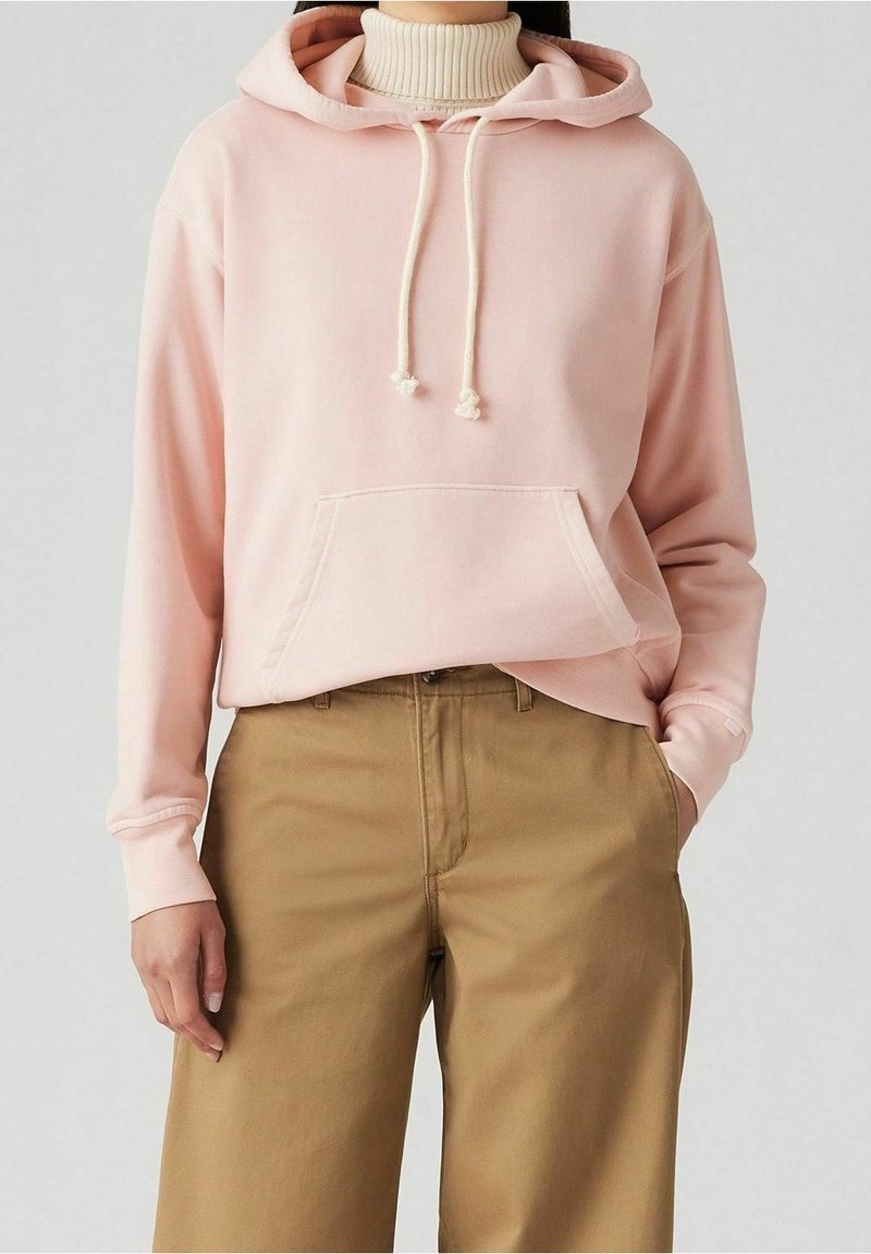 Felpa rosa chiaro con cordini regolabili, tasca frontale e polsini a coste, abbinata a pantaloni beige a gamba larga.