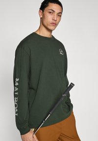 T-shirt vert à manches longues avec un motif brodé sur la poitrine et du texte imprimé sur la manche gauche. Le modèle tient un club de golf noir.