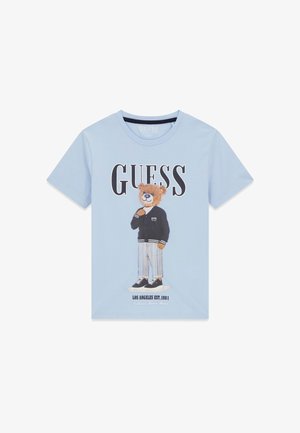 T-shirt bleu clair avec le texte "GUESS" et un ours en peluche debout portant une veste noire, un pantalon rayé et des baskets. "Los Angeles Est. 1981" en dessous.