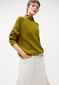 Maslinasto zelena turtleneck dukserica s teksturiranim pletenjem, dugih rukava i opuštenog kroja, uparena sa bijelim širokim hlačama.