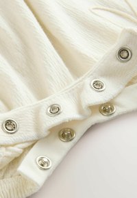 Tissu blanc à côtes avec des boutons-pression métalliques le long du bord. Présente une finition volantée et une surface texturée pour plus de détails.