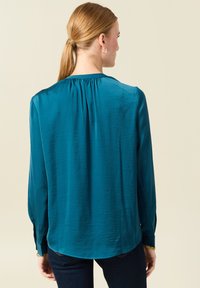 Blusa a maniche lunghe teal con un leggero lucido, dettaglio arricciato al collo sul retro e polsini con bottoni. Tessuto morbido, vestibilità comoda.