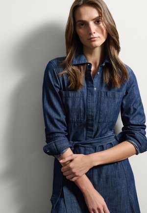 Vestido de camisa de mezclilla en azul oscuro, con cierre de botones en la parte delantera, dos bolsillos en el pecho, mangas largas y detalle de cintura atada. Textura suave.
