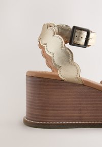 Sandale compensée avec talon empilé marron, bride de cheville métallique dorée festonnée et fermeture à boucle noire sur fond clair.