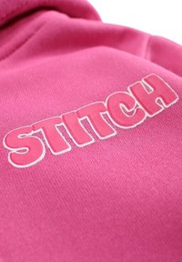 Sweat-shirt rose avec l'inscription "STITCH" en grandes lettres grasses. Les lettres ont une texture légèrement en relief avec un contour blanc.