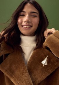 Brown faux fur coat with wide lapels over a white ribbed turtleneck, featuring a silver Christmas tree brooch on the collar.