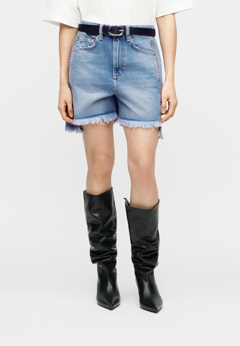 Γυναίκα που φοράει σορτς από τζιν με ψηλή μέση και ξεφτισμένα άκρα, μαύρη ζώνη και χαλαρές μαύρες δερμάτινες μπότες μέχρι το γόνατο, στέκεται μπροστά σε απλό φόντο.