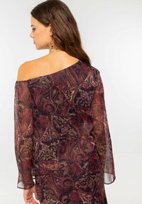 Top semitransparente sin hombros en un profundo rojo y patrón de paisley dorado. Mangas largas con puños acampanados, que cuentan con una textura suave y fluida.
