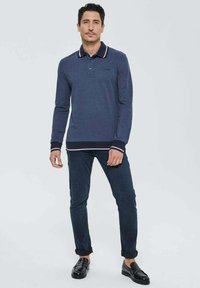 Homme se tenant debout portant une chemise polo à manches longues bleu marine avec des poignets rayés, un jean noir retroussé et des mocassins en cuir noir sur fond uni.