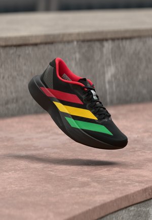 Sort atletiksko med røde, gule og grønne striber svæver over en stenovert flade udendørs.