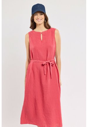 Femme portant une robe midi corail sans manches avec ceinture à la taille et casquette bleu marine, souriante sur un fond clair uni.
