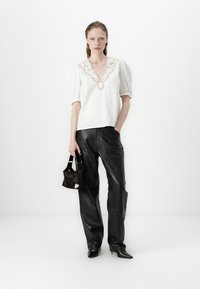 Top blanc à manches bouffantes avec un col brodé, associé à un pantalon en cuir noir. Le mannequin tient un petit sac noir avec une touche blanche.