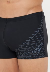 Schwarze Badehose aus elastischem Material mit einem blauen geometrischen Muster und Logo an der Seite. Enganliegendes Design mit niedrigem Bund.