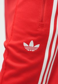 Červené sportovní kalhoty s bílými pruhy podél stran, s bílým vyšívaným logem na stehně a texturovaným vzorem látky.