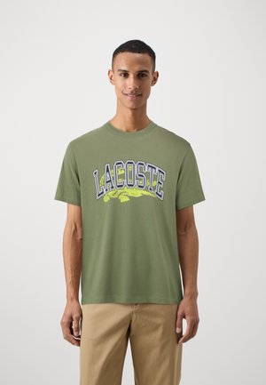 Camiseta de algodón verde con un audaz logo en azul marino y amarillo "LACOSTE" y gráfico de cocodrilo. Mangas cortas y cuello redondo.
