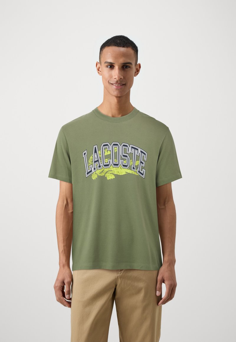 Zaļš kokvilnas T-krekls ar uzrakstu "LACOSTE" tumši zilā un dzeltenā krāsā, kā arī krokodila grafiku. Īsas piedurknes un apaļa apkakle.