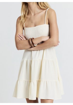 Robe à volants de couleur crème en tissu texturé avec fines bretelles spaghetti et corsage ajusté. Dotée d'une jupe évasée et fluide.