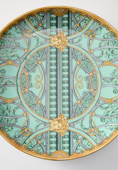 Apaļš šķīvis ar tirkīza krāsas fonu, izsmalcinātām zelta un pelēkām ziedu rakstu detaļām un centrālām seju motīvu attēlojumiem. Elegants dizains.