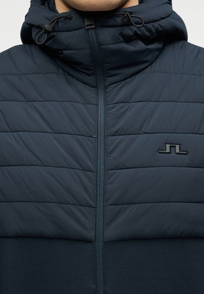 Κοντινό πλάνο ενός ατόμου που φοράει μια μπλε σκούρα μπουφάν με καπιτονέ ύφασμα, εμπρόσθιο φερμουάρ, κουκούλα με κορδόνια και ένα μικρό μαύρο λογότυπο στο στήθος.