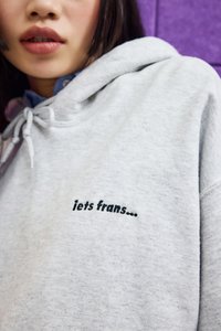 Primo piano di una persona che indossa un cappuccio grigio chiaro con il testo nero "iets frans..." ricamato sul petto, contro uno sfondo viola.