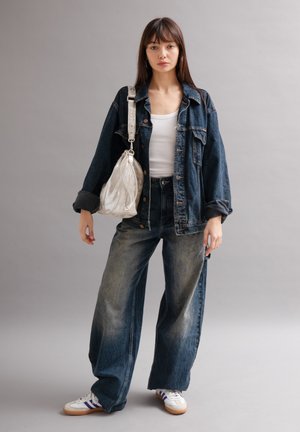 Jeune femme debout en veste en jean et jean large, débardeur blanc, baskets, portant un sac à bandoulière argenté métallique sur fond gris.