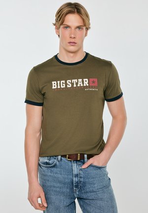 Młody mężczyzna o blond włosach, ubrany w oliwkowozielony t-shirt "Big Star" i jasnoniebieskie jeansy, z ręką w kieszeni, stojący na jednolitym tle.