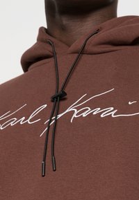 Brun hoodie i mjukt material med dragsko i huvan. Har vit broderad text "Karl" på framsidan, vilket framhäver unika design detaljer.