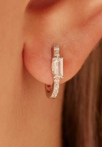 Zilveren oorring met een rechthoekige duidelijke edelsteen omringd door kleinere kristallen, met een glanzende metalen afwerking en een slank ontwerp.