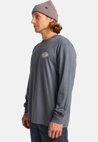 Langärmliges graues T-Shirt aus Baumwolle, mit einem runden Halsausschnitt und einem gestickten Logo auf der linken Brust. Getragen mit einer helllila Mütze.
