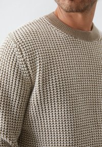 Maglione lavorato a maglia di colore beige con un motivo testurizzato, caratterizzato da un collo a coste. Il design include sottili variazioni di colore.