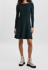 Femme portant une robe longue à manches longues de couleur vert foncé, arrivant au genou, associée à des bottines à lacets beige clair avec des semelles noires épaisses.