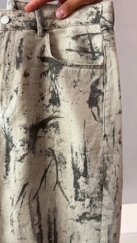 Mano che tiene dei jeans grigi con motivi di schizzi di vernice nera astratti vicino alla tasca anteriore e al bottone della cintura.