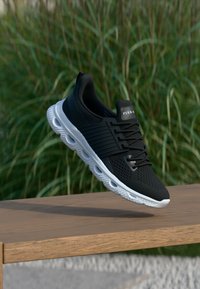 Μαύρο αθλητικό παπούτσι με επάνω μέρος από δίχτυ, υφασμένα διακοσμητικά στοιχεία και λευκή σόλα με χαρακτηριστικό κυματιστό σχέδιο. Τα κορδόνια είναι μαύρα, το λογότυπο βρίσκεται στη γλώσσα του παπουτσιού.