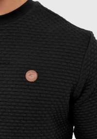 Suéter negro texturizado con un cuello acanalado y un parche de cuero redondo en el pecho. El diseño resalta un patrón tejido.