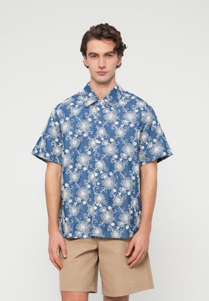 Giovane uomo che indossa una camicia floreale a maniche corte blu e pantaloncini beige, in piedi davanti a uno sfondo grigio chiaro uniforme.