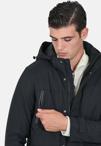 Giacca nera imbottita con tasca con cerniera sul petto, chiusure a pressione e cappuccio. Polsini strutturati completano il design.