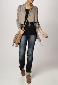 Wrap in khaki con frange, top nero, jeans slim in denim, stivaletti marroni alla caviglia. Materiale morbido, vestibilità rilassata, borsa testurizzata e leggeri dettagli drappeggiati.