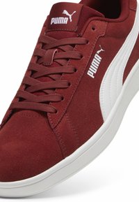 Bordowe zamszowe sneakersy Puma z białym logo i sznurówkami w tym samym kolorze, z białą gumową podeszwą, ustawione pod kątem, aby pokazać szczegóły z boku i z góry.