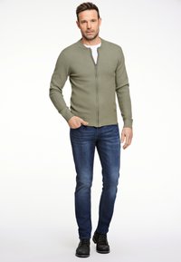 Maglione zip-up verde oliva a maglia con scollatura rotonda, abbinato a jeans aderenti blu scuro e stivali neri. Design semplice e strutturato.