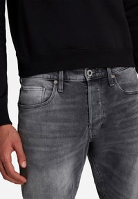 Grå denimjeans med femficksdesign, dragkedja och subtila blekningar. Svart sweatshirt med ribbad nederkant synlig ovanför jeansen.